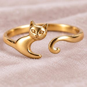 Op de afbeelding: Een goudkleurige open ring met een kattenontwerp. De ringband is gevormd als het lichaam van een kat, met de kop aan het ene uiteinde en een gekrulde staart aan het andere uiteinde. Het oppervlak van de ring is glad en gepolijst.