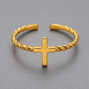 May include: A gold-toned ring featuring a polished cross centrepiece attached to a twisted rope-style band. The ring has an open design at the back, allowing for adjustable sizing. The metal surface has a smooth, reflective finish throughout.