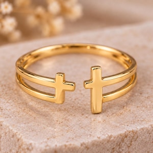 Peut inclure: Une bague ouverte dorée présentant deux petits motifs en forme de croix aux extrémités de l'anneau. La bague possède une finition lisse et polie ainsi qu'un design à double anneau relié à chaque croix, créant un espace entre les deux symboles.
