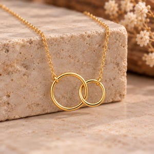 Könnte beinhalten: Eine goldfarbene Halskette mit zwei ineinandergreifenden, unterschiedlich großen kreisförmigen Ringen, die an einer feinen Kette hängen. Das Metall hat eine polierte, reflektierende Oberfläche und das Design besteht aus einem größeren Ring.