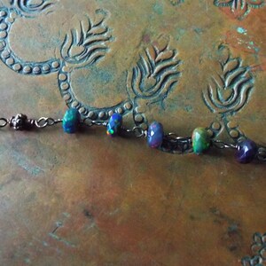 May include: A delicate silver chain necklace with iridescent opal and amethyst beads. The necklace is displayed on a copper surface with an ornate design.