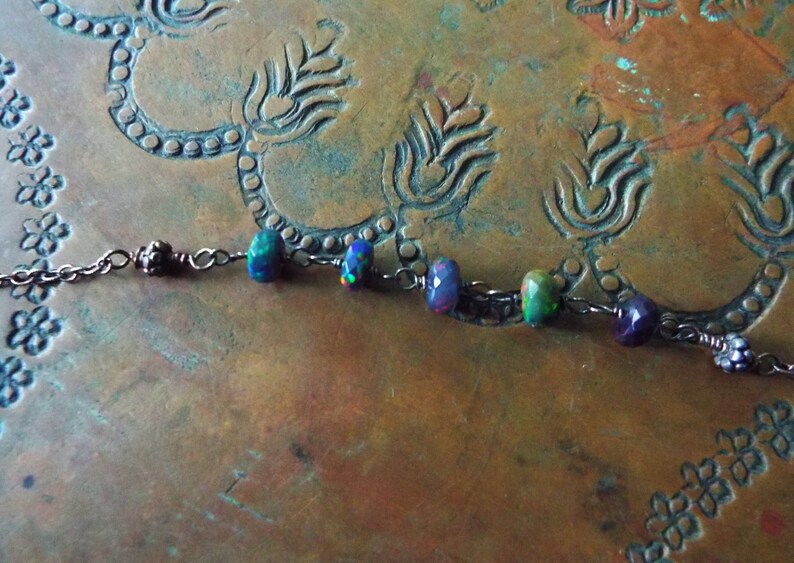 May include: A delicate silver chain necklace with iridescent opal and amethyst beads. The necklace is displayed on a copper surface with an ornate design.