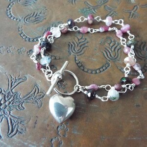 Pode incluir: Um bracelete de prata com um fecho de cora&ccedil;&atilde;o e um fecho de correr. O bracelete &eacute; feito com uma dupla corrente de prata e apresenta pedras preciosas rosa, verdes e azuis.