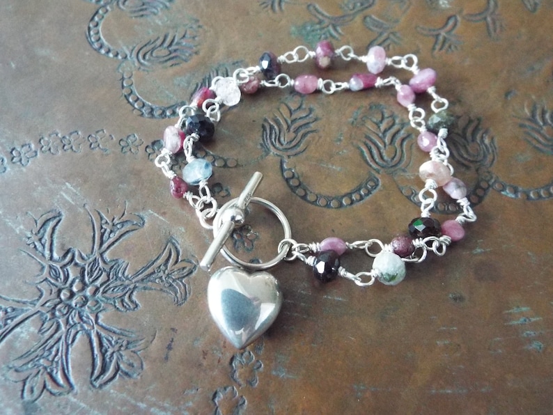 Pode incluir: Um bracelete de prata com um fecho de cora&ccedil;&atilde;o e um fecho de correr. O bracelete &eacute; feito com uma dupla corrente de prata e apresenta pedras preciosas rosa, verdes e azuis.