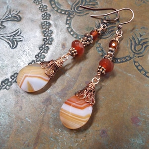 以下が含まれることがあります： アゲートの涙型ストーンとカーネリアンのビーズが付いた銅製のイヤリング。イヤリングはフィリグリーのデザインで、暖かくアースカラーです。