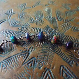 May include: A close-up of a delicate necklace featuring a dark metal chain and a series of iridescent opal beads. The beads display a mix of dark blue, purple, and red tones, with a hint of green.