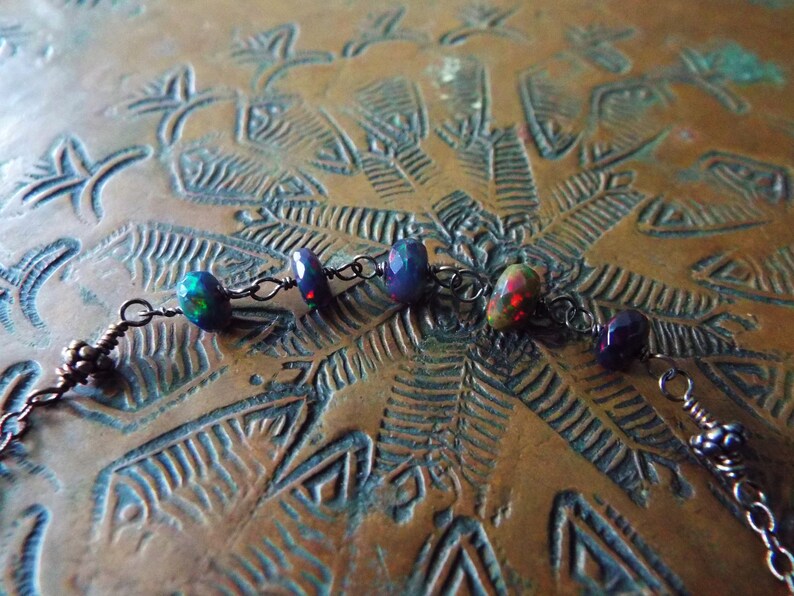 May include: A close-up of a delicate necklace featuring a dark metal chain and a series of iridescent opal beads. The beads display a mix of dark blue, purple, and red tones, with a hint of green.