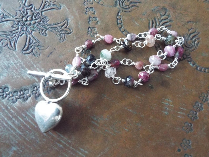 Pode incluir: Um bracelete de corrente de prata com um fecho de cora&ccedil;&atilde;o e pedras preciosas multicoloridas. O bracelete tem um fecho de correr.