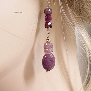 May include: A gold-colored earring with a series of purple and pink gemstones. The earring features a hook, a cluster of small, round stones, a set of faceted gems, a stacked section, and a large oval stone. The text "Rust & Salt" is visible.