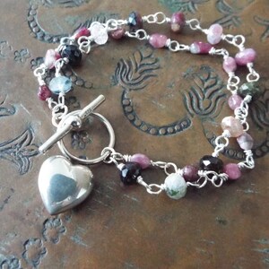 Pode incluir: Um bracelete de prata com um fecho de cora&ccedil;&atilde;o e um fecho de correr. O bracelete &eacute; feito com uma dupla corrente de prata e apresenta pedras preciosas rosa, verdes e pretas.