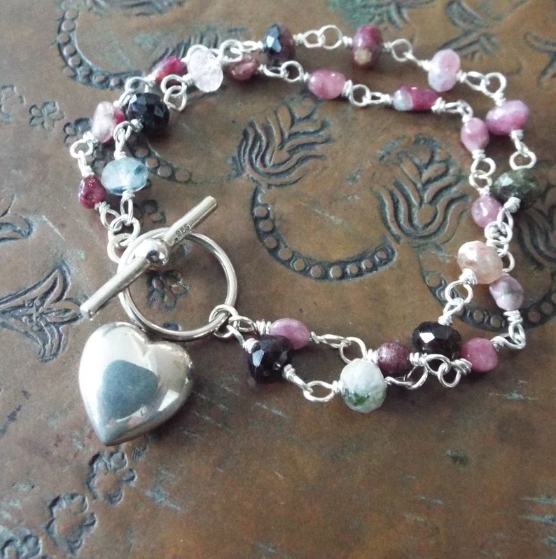 Pode incluir: Um bracelete de prata com um fecho de cora&ccedil;&atilde;o e um fecho de correr. O bracelete &eacute; feito com uma dupla corrente de prata e apresenta pedras preciosas rosa, verdes e pretas.