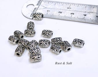 Pode incluir: Um close-up de um monte de contas de metal prateado com um design ornamentado. As contas medem aproximadamente 2,5 cm de comprimento e têm um furo no centro. As contas estão sobre uma superfície branca com uma régua no fundo. A régua está marcada em polegadas e centímetros. O texto "Rust & Salt" é visível no canto inferior direito da imagem.