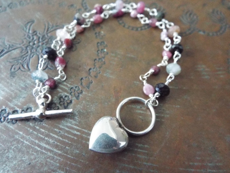 Pode incluir: Um colar de corrente de prata com um pingente de cora&ccedil;&atilde;o e um fecho de correr. A corrente &eacute; adornada com contas rosa, verde e pretas.