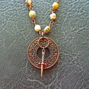 May include: A copper necklace with a round pendant featuring a decorative design and a small dagger hanging from it. The necklace is made with brown and yellow beads.