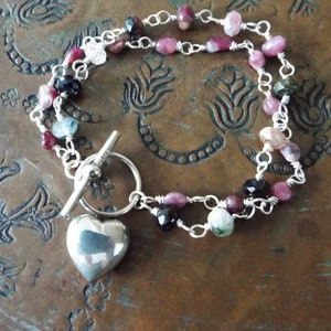 Pode incluir: Um bracelete de prata com um fecho de cora&ccedil;&atilde;o e um fecho de correr. O bracelete &eacute; feito com uma dupla corrente de prata e apresenta pedras preciosas rosa, verdes e pretas.