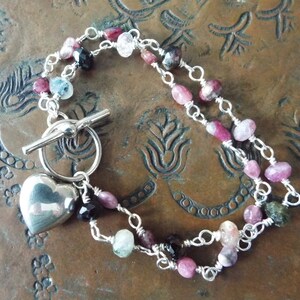 Pode incluir: Um bracelete de prata com um fecho de cora&ccedil;&atilde;o e um fecho de correr. O bracelete apresenta pedras preciosas rosa, verde e preta.