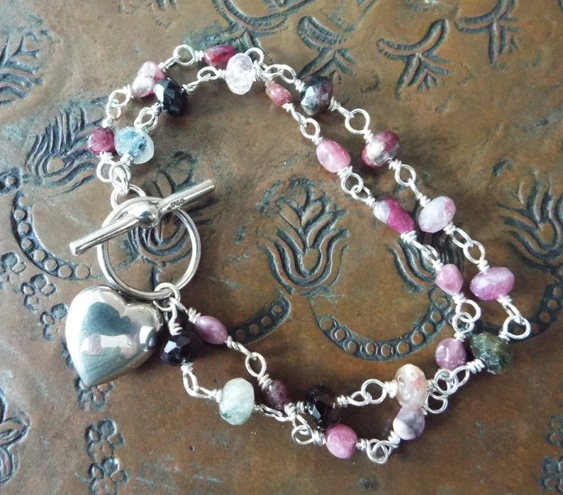 Pode incluir: Um bracelete de prata com um fecho de cora&ccedil;&atilde;o e um fecho de correr. O bracelete apresenta pedras preciosas rosa, verde e preta.