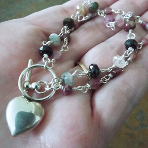 Pode incluir: Um colar de prata com um pingente em forma de cora&ccedil;&atilde;o e um fecho de correr. O colar apresenta uma dupla corrente de fio com v&aacute;rias pedras preciosas coloridas, incluindo rosa, verde, preto e transparente.