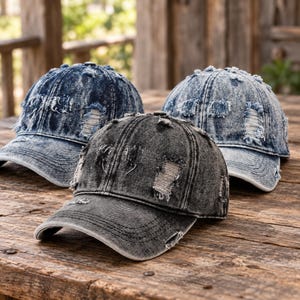 May include: Three baseball caps made of distressed denim fabric. Two caps feature a light blue acid-wash finish, while the central cap is in a dark charcoal grey acid-wash. Each hat displays frayed, ripped detailing across the crown and brim.