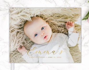 以下が含まれることがあります： 「Emma's First Birthday - June 18, 2022」と金色の文字で書かれた白い写真アルバム。アルバムには、ふわふわの白い毛布の上に横になっている赤ちゃんの写真が載っています。