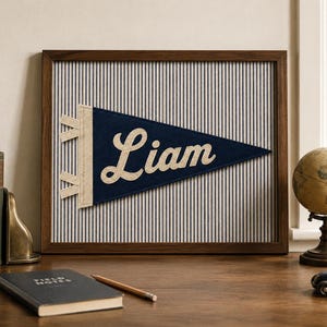 Puede incluir: Un cuadro decorativo que presenta un banderín de fieltro azul oscuro con el nombre Liam en letras cursivas de color crema. El banderín está montado sobre un fondo de tela blanca con finas rayas verticales azul marino, dentro de un marco de madera.