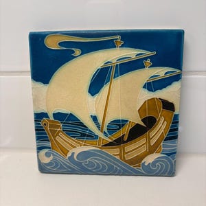 May include: A square ceramic tile featuring a relief illustration of a wooden sailing ship on a deep blue sea. The ship has cream-coloured sails and a golden-brown hull, navigating through stylised blue waves under a dark blue sky with clouds.