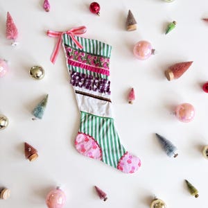 Op de afbeelding: Een kerstkous met een patchworkontwerp van groene en witte verticale strepen, roze bloemenborduursels, paarse gingham ruches en een roze patroon bij de teen en hiel. Een roze en wit gestreepte strik is aan de bovenhoek bevestigd.