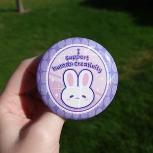 Könnte beinhalten: Ein runder Ansteckbutton mit violettem Wellenrand und holografischer Oberfläche. In der Mitte ist ein weißes Cartoon-Hasengesicht mit rosa Ohren sowie der Text I support human creativity in violetter Schrift auf hellem Grund zu sehen.