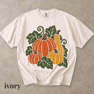 May include: An ivory short-sleeve t-shirt featuring a central graphic print of three pumpkins in shades of orange and yellow, surrounded by dark green leaves and vines. The shirt has a crew neckline and a visible brand tag at the inner collar.