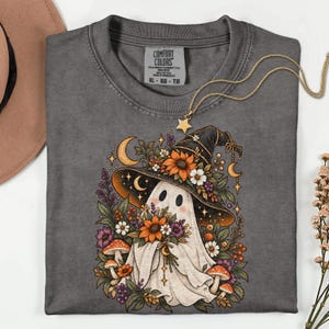 May include: A charcoal gray t-shirt featuring a graphic of a white ghost wearing a black witch hat adorned with sunflowers, daisies, and foliage. The design includes crescent moons, stars, and mushrooms, with the Comfort Colors brand label visible.