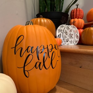 May include: A large orange artificial pumpkin with the text happy fall written in black cursive script. The pumpkin is displayed alongside several other smaller orange pumpkins and a white decorative pumpkin with a leaf cutout pattern.