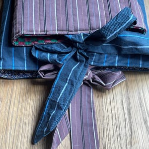 Può includere: Un involucro trapuntato caratterizzato da un tessuto color malva con strisce verticali bianche e nere sovrapposto a un tessuto blu con motivo a griglia. L'involucro è fissato con lunghi lacci in tessuto a righe malva e a quadretti blu, rivelando una fodera a scacchi verdi e rossi.