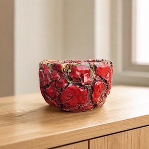 May include: A small, round ceramic bowl with a vibrant red, crackled glaze finish. The surface features deep, irregular fissures that reveal dark, textured crevices and subtle gold-toned accents throughout the exterior of the vessel.