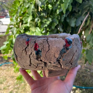 以下が含まれることがあります： ひび割れたような質感の茶色の表面を持つ、手作りの丸い陶器製プランター。外側には不規則な亀裂があり、光沢のある濃い赤と青緑色の釉薬が垂れたアクセントになっています。鉢は小さなブロック状の脚の上に置かれています。