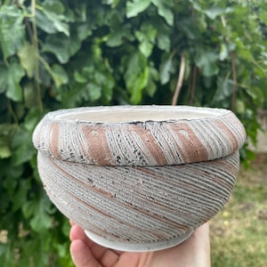 May include: A round ceramic bowl featuring a textured exterior with diagonal grooves in alternating shades of terracotta orange and weathered gray. The rim is thick and rounded, while the interior surface appears smooth and off-white in color.