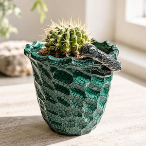 May include: A dark green ceramic planter with a textured, crackled hexagonal pattern and a wavy rim. A small, sculpted dark gray snake figure is attached to the upper edge of the pot. A round, spiky green cactus is planted inside the container.