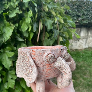 Può includere: Un vaso in ceramica a forma di elefante con una smaltatura testurizzata e maculata rosa e bianca. Il contenitore presenta grandi orecchie piatte, occhi rotondi prominenti e una proboscide curva sul davanti con una finitura screpolata.