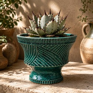 Op de afbeelding: Een blauwgroene keramische plantenbak met een uitlopende rand en een textuur met ribbelpatroon. De pot heeft een glanzende, craquelé glazuurafwerking en staat op een voetstuk. Een stekelige, lichtgroene agave vetplant staat in de pot.