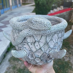May include: A round, off-white planter featuring a textured, scaly surface pattern resembling reptile skin. A sculpted snake wraps around the upper rim, while four small, pointed, scaly appendages extend from the sides of the vessel body.