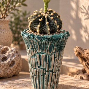 Op de afbeelding: Een donkergroene, geribbelde cactus met een enkele witte bloem die bovenop bloeit, geplant in een getextureerde, blauwgroene en zilveren keramische pot met een ruw, verticaal patroon en een grillige, onregelmatige rand.