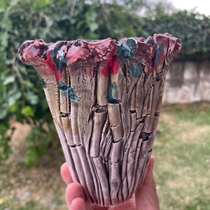 May include: A ceramic planter with a textured, bark-like surface in shades of beige and grey. The rim features a thick, uneven glaze in deep red and dark teal, with vertical fissures running down the body of the vessel, creating a rugged appearance.