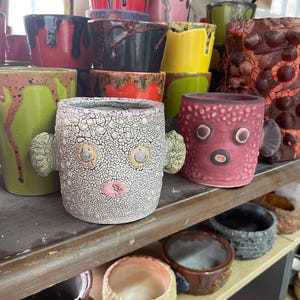 Puede incluir: Una colección de tazas y macetas de cerámica en varios colores y texturas. Dos tazas destacadas en primer plano presentan rostros esculpidos con protuberancias laterales, una con esmalte craquelado blanco y otra en acabado rosa mate.