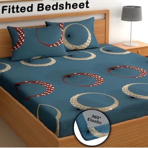 Op de afbeelding: Een blauw hoeslaken en bijpassende kussensloop met een herhalend patroon van grote cirkels in bruin- en beige tinten. De tekst Fitted Bedsheet staat bovenaan en een cirkelvormig inzetstuk toont 360 Elastic.