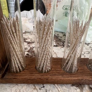 May include: A wooden stand featuring three vertical glass cylinders, each filled with a collection of slender, silver-toned, spiral-textured sticks. The base is crafted from dark brown wood, providing a stable foundation for the glass tubes.