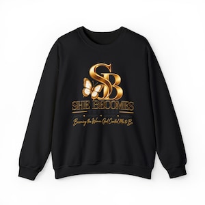 May include: A black long-sleeve sweatshirt featuring a gold metallic graphic on the chest. The design includes a large stylized SB monogram with a butterfly, the text SHE BECOMES THE BLOG, and the phrase Becoming the Woman God Created Me to Be.