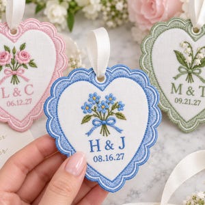 May include: Three heart-shaped fabric ornaments with scalloped embroidered edges and white satin ribbons. Each features a floral bouquet, initials, and a date. The pink ornament shows roses, the blue shows forget-me-nots, and the green shows lily of the valley.