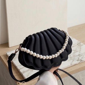 Peut inclure: Un sac à main noir en forme de coquillage avec un design incurvé et nervuré, orné d'un collier de perles blanches sur le devant. Le sac possède une fine bandoulière noire avec des détails dorés et le texte ANDRA écrit en lettres dorées.