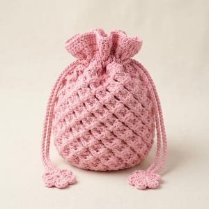 Op de afbeelding: Een lichtroze gehaakt trekkoordtasje met een gestructureerd ruitpatroon over het hele lichaam. De tas heeft een geplooide opening met twee lange, bijpassende roze koorden die eindigen in kleine, zesbladige bloemvormige gehaakte accenten.