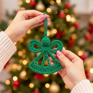 May include: A green crocheted angel ornament featuring a circular head, two symmetrical open-work wings, and a flared skirt base with a hanging loop at the top. The ornament is crafted from yarn with a visible stitch texture throughout.