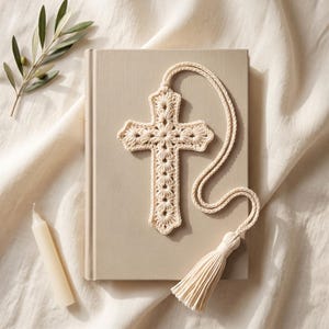 May include: A cream-colored crocheted cross ornament featuring a delicate floral-inspired stitch pattern. The cross is attached to a matching braided cord that ends in a long, thick tassel, creating a cohesive and textured decorative piece.
